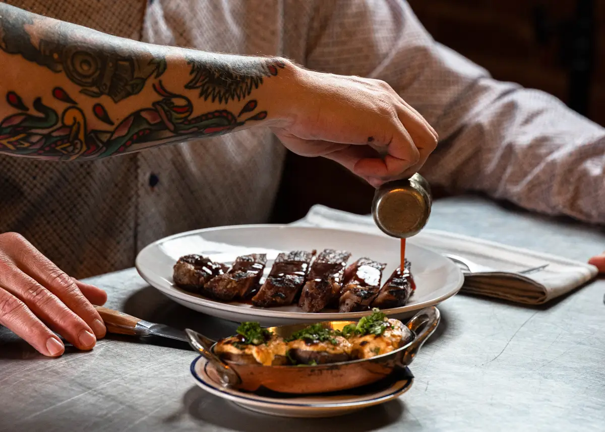 a tattooed arm pours jus on a prepared steak at freight house